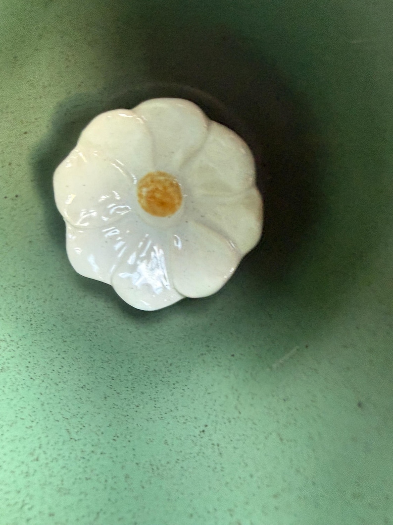 May include: A white ceramic flower-shaped lid with a yellow center. The lid is set against a green background, creating a contrast. The flower design adds a touch of elegance.