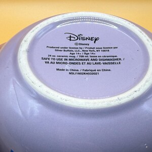May include: A lavender ceramic mug with the Disney logo and text. The mug is 24 oz. and safe for microwave and dishwasher use. The text includes the manufacturer's information and the phrase "Made in China."