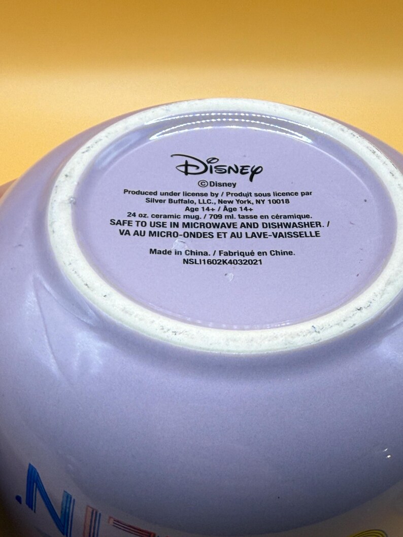 May include: A lavender ceramic mug with the Disney logo and text. The mug is 24 oz. and safe for microwave and dishwasher use. The text includes the manufacturer's information and the phrase "Made in China."