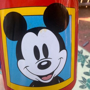 May include: A cylindrical mug with a bright red exterior featuring a central graphic of Mickey Mouse. The character is depicted in black and white with a smiling expression, set against a light blue square background framed by a thick yellow border.