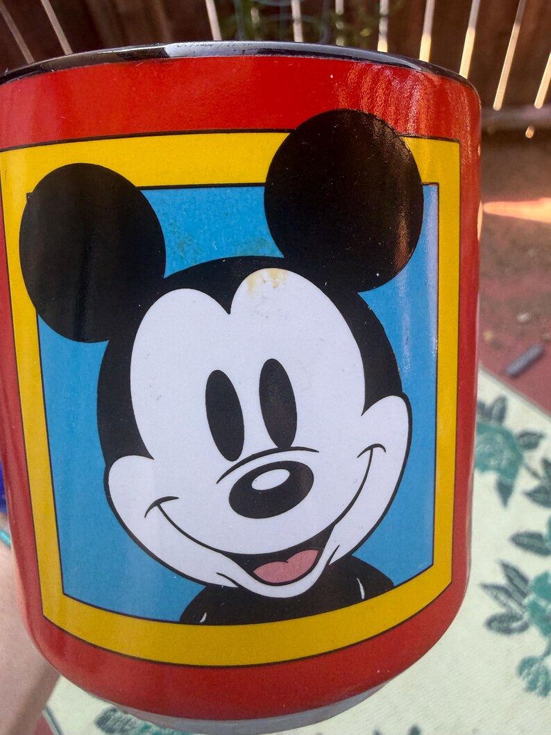May include: A cylindrical mug with a bright red exterior featuring a central graphic of Mickey Mouse. The character is depicted in black and white with a smiling expression, set against a light blue square background framed by a thick yellow border.