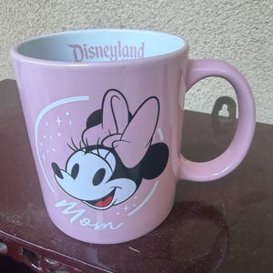 May include: A light pink ceramic mug featuring a graphic of Minnie Mouse with a pink bow. The word Mom is printed in white script below the character. The interior rim of the mug displays the word Disneyland in pink text against a white background.