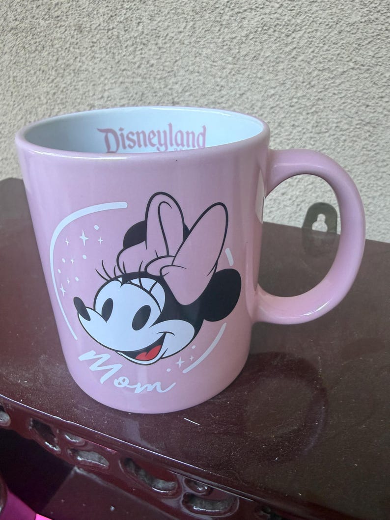 May include: A light pink ceramic mug featuring a graphic of Minnie Mouse with a pink bow. The word Mom is printed in white script below the character. The interior rim of the mug displays the word Disneyland in pink text against a white background.