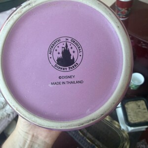 Puede incluir: La base circular de una taza de cer&aacute;mica con una superficie de color morado claro y un logotipo impreso en negro. El logotipo muestra la silueta de un castillo rodeado de estrellas y el texto AUTHENTIC ORIGINAL DISNEY PARKS. Debajo, DISNEY y MADE IN THAILAND.