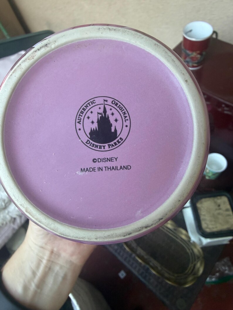 Puede incluir: La base circular de una taza de cer&aacute;mica con una superficie de color morado claro y un logotipo impreso en negro. El logotipo muestra la silueta de un castillo rodeado de estrellas y el texto AUTHENTIC ORIGINAL DISNEY PARKS. Debajo, DISNEY y MADE IN THAILAND.