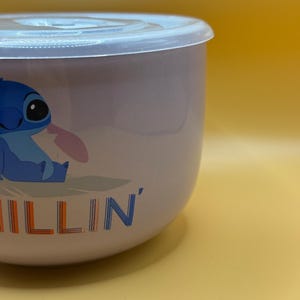 May include: A light purple bowl with a clear lid. The bowl features a blue cartoon character with the word "ILLIN'" in orange and blue. The bowl is on a yellow background.