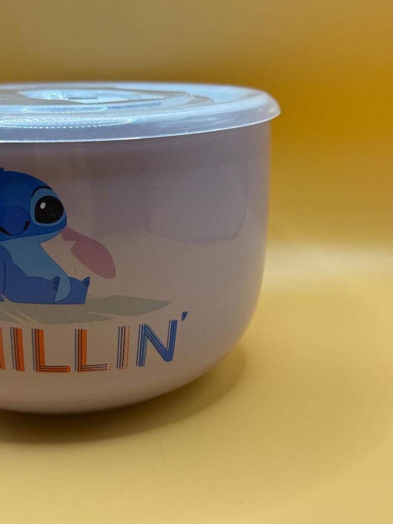 May include: A light purple bowl with a clear lid. The bowl features a blue cartoon character with the word "ILLIN'" in orange and blue. The bowl is on a yellow background.