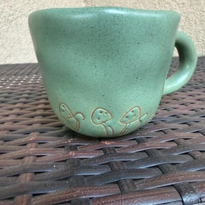 May include: A green ceramic mug with a speckled finish and a curved handle. The mug features a series of embossed mushroom designs around the base. The mug is sitting on a brown woven surface.