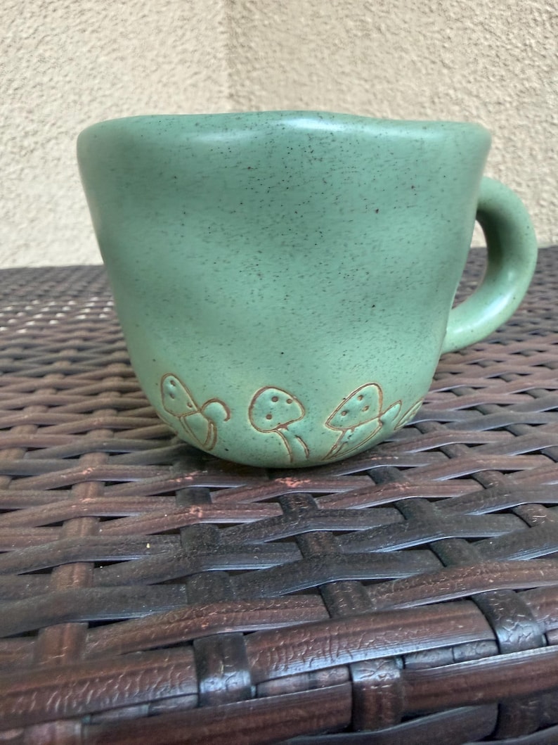 May include: A green ceramic mug with a speckled finish and a curved handle. The mug features a series of embossed mushroom designs around the base. The mug is sitting on a brown woven surface.