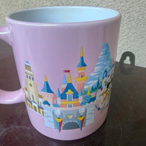 May include: A light pink ceramic mug featuring a colorful graphic illustration of a castle with blue and yellow turrets, a mountain peak, and a circular ride structure. The interior of the mug is white, and it has a curved handle on the left side.