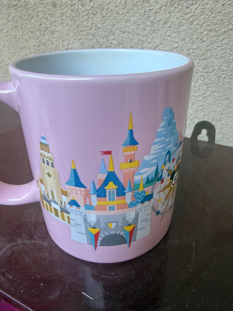 May include: A light pink ceramic mug featuring a colorful graphic illustration of a castle with blue and yellow turrets, a mountain peak, and a circular ride structure. The interior of the mug is white, and it has a curved handle on the left side.