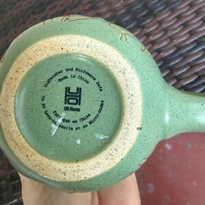 May include: A light green ceramic mug with a handle, featuring a speckled texture and a beige rim. The bottom of the mug has text that reads "Dishwasher and Microwave Safe Made in China". The mug has a unique design.
