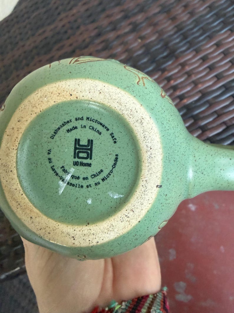 May include: A light green ceramic mug with a handle, featuring a speckled texture and a beige rim. The bottom of the mug has text that reads "Dishwasher and Microwave Safe Made in China". The mug has a unique design.