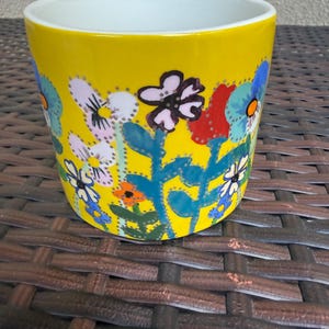 May include: A yellow ceramic flower pot with a white interior. The exterior is decorated with colorful hand-painted flowers in shades of blue, red, orange, and white. The pot sits on a brown woven surface.