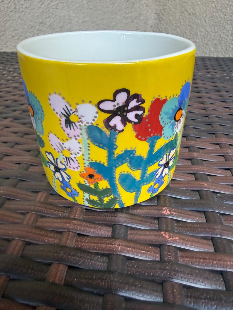 May include: A yellow ceramic flower pot with a white interior. The exterior is decorated with colorful hand-painted flowers in shades of blue, red, orange, and white. The pot sits on a brown woven surface.