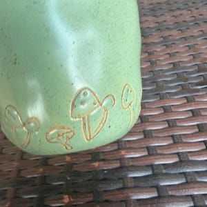 May include: A light green ceramic mug with a textured surface. The mug features a series of etched mushroom designs in a lighter brown color. The mug is partially visible, showcasing its rounded shape and the detailed mushroom illustrations.