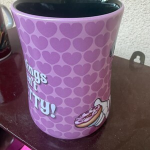 Puede incluir: Una taza de cer&aacute;mica con interior negro y exterior de color morado claro con un patr&oacute;n repetitivo de corazones peque&ntilde;os. La taza muestra un gr&aacute;fico de una mano de dibujos animados sosteniendo un donut y el texto blanco parcial "ings n't TY!"