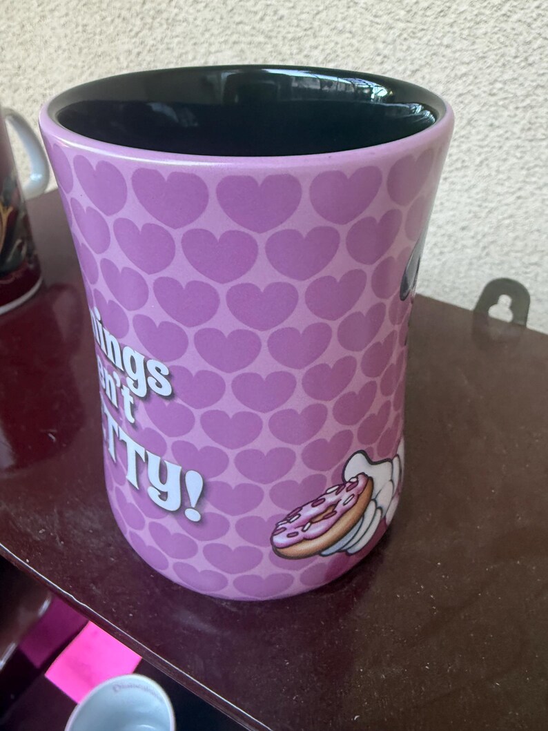 Puede incluir: Una taza de cer&aacute;mica con interior negro y exterior de color morado claro con un patr&oacute;n repetitivo de corazones peque&ntilde;os. La taza muestra un gr&aacute;fico de una mano de dibujos animados sosteniendo un donut y el texto blanco parcial "ings n't TY!"
