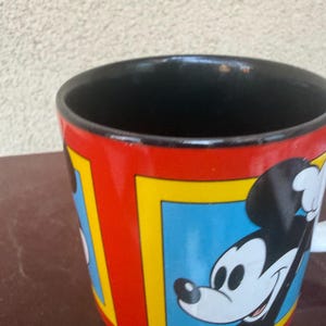 May include: A ceramic mug with a glossy red exterior featuring multiple rectangular frames with yellow borders. Each frame contains a blue background and a graphic illustration of Mickey Mouse. The interior of the mug is solid black.