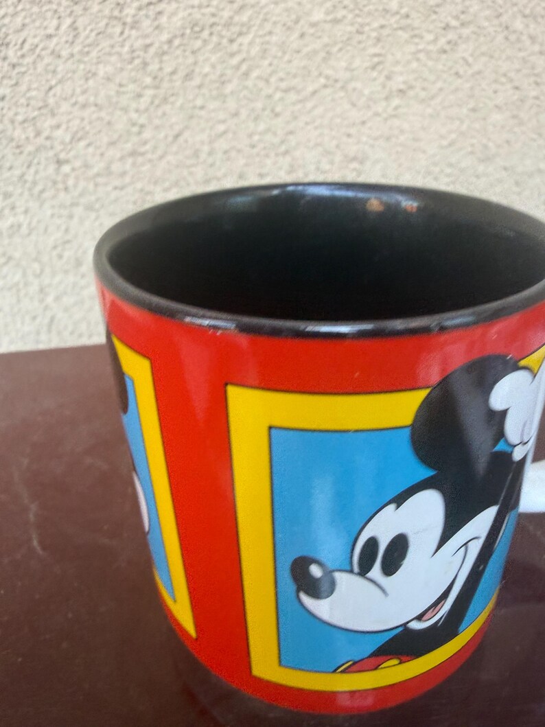 May include: A ceramic mug with a glossy red exterior featuring multiple rectangular frames with yellow borders. Each frame contains a blue background and a graphic illustration of Mickey Mouse. The interior of the mug is solid black.