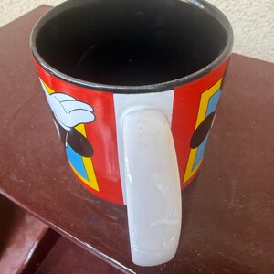 May include: A ceramic mug with a black interior and a white handle. The exterior features a red background with vertical white stripes and rectangular panels containing blue borders, displaying cartoon character graphics with white gloved hands.