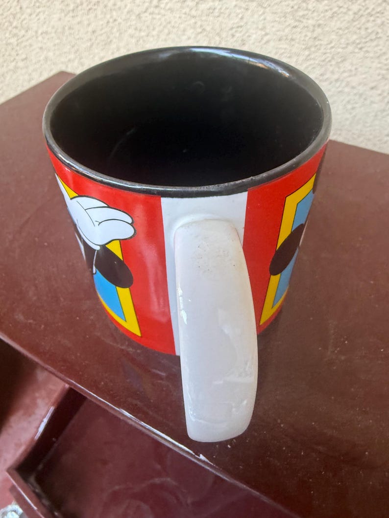 May include: A ceramic mug with a black interior and a white handle. The exterior features a red background with vertical white stripes and rectangular panels containing blue borders, displaying cartoon character graphics with white gloved hands.
