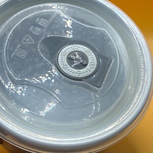 May include: A close-up of a white and clear plastic container lid. The lid has a clear plastic center with a dial that reads from 5 to 30. The lid also has a series of symbols and text printed on it.
