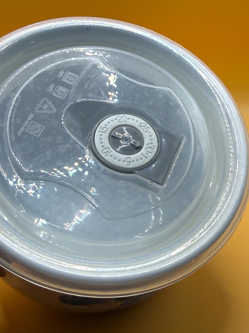May include: A close-up of a white and clear plastic container lid. The lid has a clear plastic center with a dial that reads from 5 to 30. The lid also has a series of symbols and text printed on it.