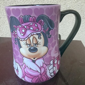 Puede incluir: Una taza de cer&aacute;mica con fondo rosa y un patr&oacute;n repetitivo de corazones. El frente muestra un gr&aacute;fico de Minnie Mouse con un antifaz rosa de lunares y una bata rosa con ribete de piel blanca. La taza tiene un asa y un interior de color negro.