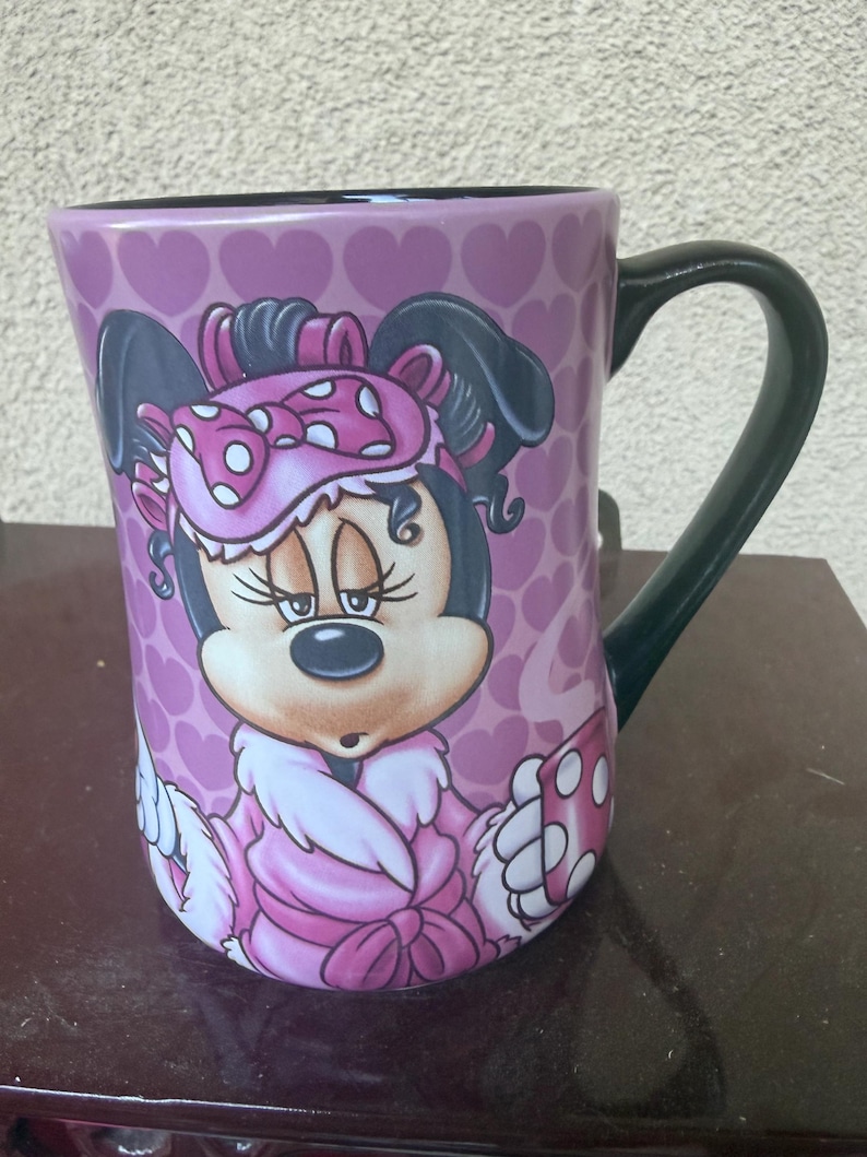 Puede incluir: Una taza de cer&aacute;mica con fondo rosa y un patr&oacute;n repetitivo de corazones. El frente muestra un gr&aacute;fico de Minnie Mouse con un antifaz rosa de lunares y una bata rosa con ribete de piel blanca. La taza tiene un asa y un interior de color negro.