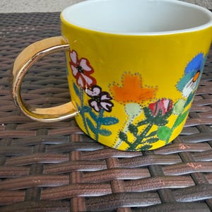 May include: A yellow ceramic mug with a gold handle and a white interior. The mug is decorated with colorful floral designs in red, pink, orange, and blue. The mug is sitting on a brown woven surface.