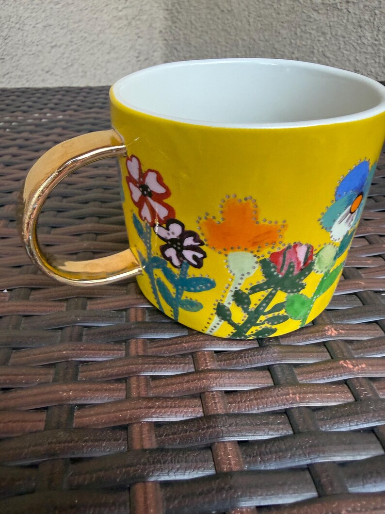 May include: A yellow ceramic mug with a gold handle and a white interior. The mug is decorated with colorful floral designs in red, pink, orange, and blue. The mug is sitting on a brown woven surface.