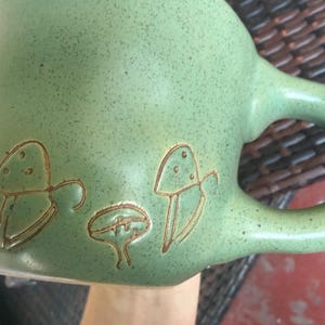 May include: A green ceramic mug with a speckled finish and a large handle. The mug features brown etched mushroom designs on the side. The mug is held by a person.
