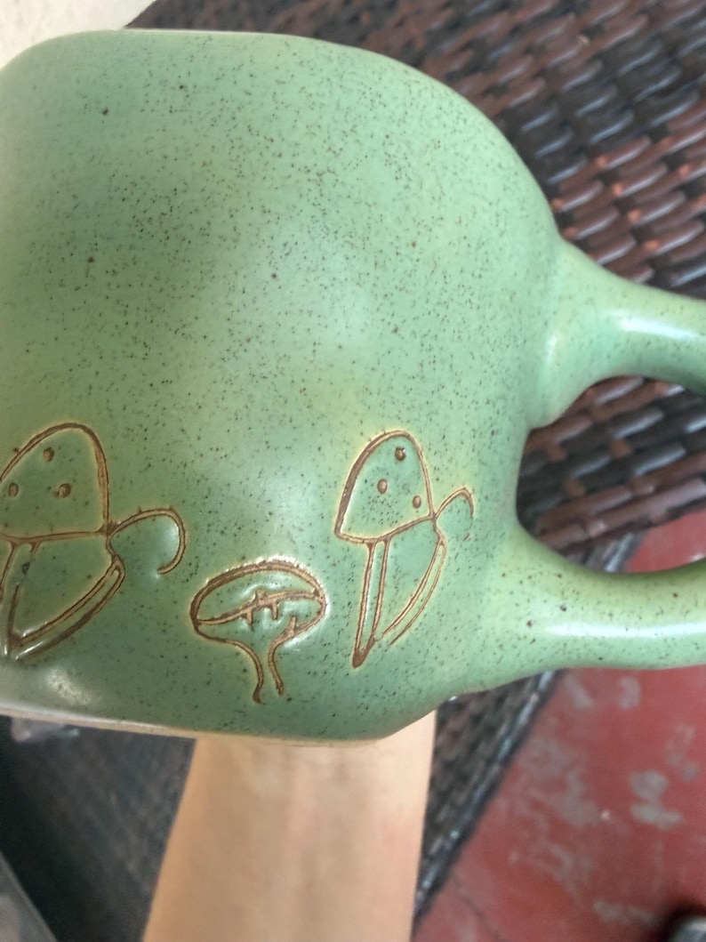 May include: A green ceramic mug with a speckled finish and a large handle. The mug features brown etched mushroom designs on the side. The mug is held by a person.