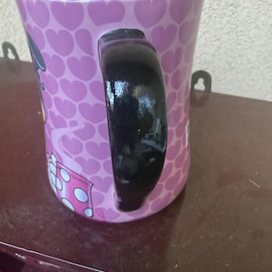 Puede incluir: Una taza de cer&aacute;mica con interior y asa de color negro brillante. El exterior presenta un fondo de color p&uacute;rpura claro cubierto con un patr&oacute;n repetitivo de corazones de color p&uacute;rpura oscuro. Se ve parte de un gr&aacute;fico con una caja de lunares.
