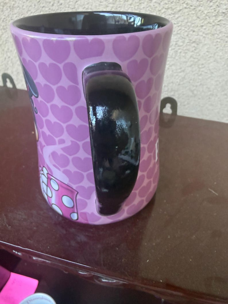 Puede incluir: Una taza de cer&aacute;mica con interior y asa de color negro brillante. El exterior presenta un fondo de color p&uacute;rpura claro cubierto con un patr&oacute;n repetitivo de corazones de color p&uacute;rpura oscuro. Se ve parte de un gr&aacute;fico con una caja de lunares.