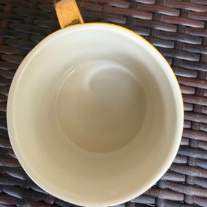 May include: A yellow and cream-colored ceramic mug with a wooden handle. The mug's interior is cream-colored, and the exterior rim is yellow. The handle is made of wood and is attached to the side of the mug.
