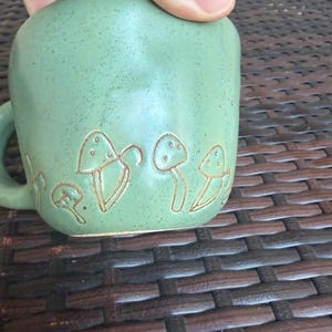May include: A green ceramic mug with a handle, featuring an embossed mushroom design. The mug has a speckled texture and a rounded shape. The mushroom design is in a lighter shade of green, creating a contrast. The mug is held by a hand.