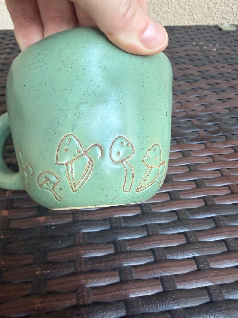 May include: A green ceramic mug with a handle, featuring an embossed mushroom design. The mug has a speckled texture and a rounded shape. The mushroom design is in a lighter shade of green, creating a contrast. The mug is held by a hand.