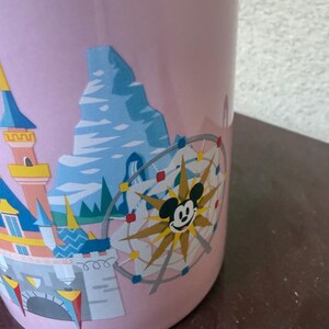 May include: A light pink ceramic mug featuring a colorful graphic print of a castle, a mountain peak, and a circular Ferris wheel ride with a central Mickey Mouse icon. The design includes blue, yellow, and red accents against the pastel background.