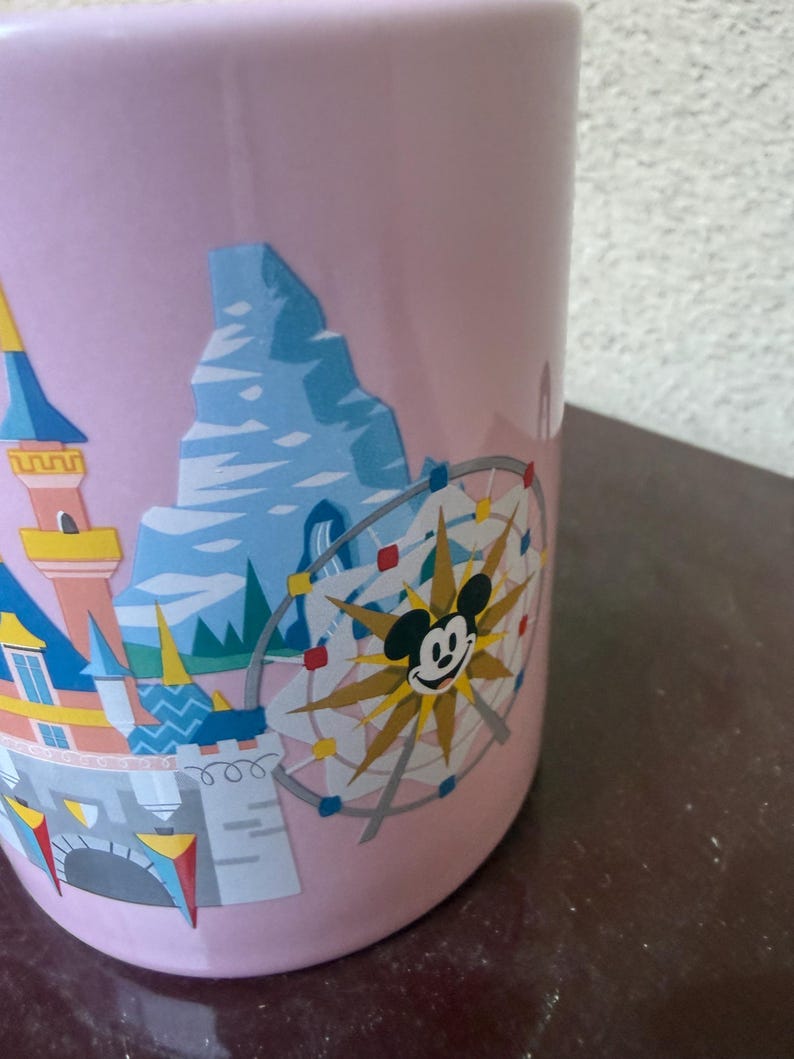 May include: A light pink ceramic mug featuring a colorful graphic print of a castle, a mountain peak, and a circular Ferris wheel ride with a central Mickey Mouse icon. The design includes blue, yellow, and red accents against the pastel background.