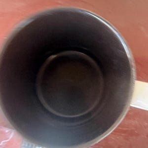 May include: A top-down view of a cylindrical mug featuring a solid black interior and a white exterior surface. The mug has a smooth, rounded rim and a single handle extending from the side, captured from a high angle looking directly into the vessel.