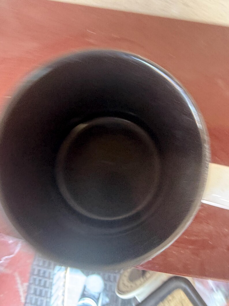 May include: A top-down view of a cylindrical mug featuring a solid black interior and a white exterior surface. The mug has a smooth, rounded rim and a single handle extending from the side, captured from a high angle looking directly into the vessel.