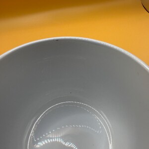 May include: A white ceramic bowl with a smooth, glossy finish. The bowl has a rounded shape and a wide rim. The interior of the bowl reflects light, creating a subtle sheen. The background is a solid orange color.