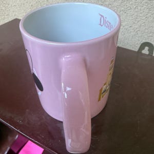 May include: A light pink ceramic mug with a glossy finish and a large handle. The interior is white and features the word Disney printed in pink script near the rim. The exterior displays a graphic of a castle and a black and white character silhouette.