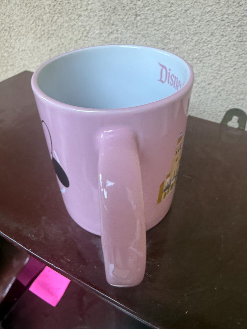 May include: A light pink ceramic mug with a glossy finish and a large handle. The interior is white and features the word Disney printed in pink script near the rim. The exterior displays a graphic of a castle and a black and white character silhouette.