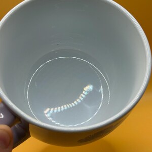 May include: A white ceramic mug with a light purple handle. The interior of the mug is a light blue color. The mug is set against a yellow background.