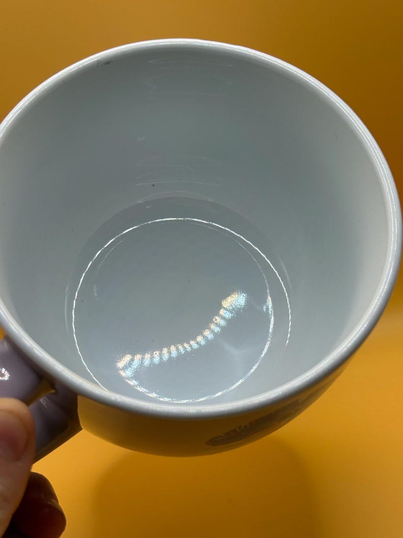 May include: A white ceramic mug with a light purple handle. The interior of the mug is a light blue color. The mug is set against a yellow background.