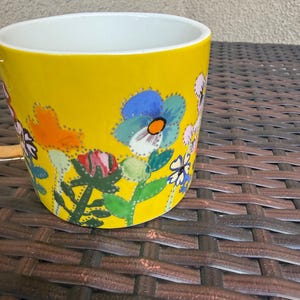 May include: A yellow ceramic mug with a white interior, adorned with a colorful floral design. The flowers include blue, orange, pink, and white blooms with green leaves. The mug is cylindrical and sits on a brown woven surface.