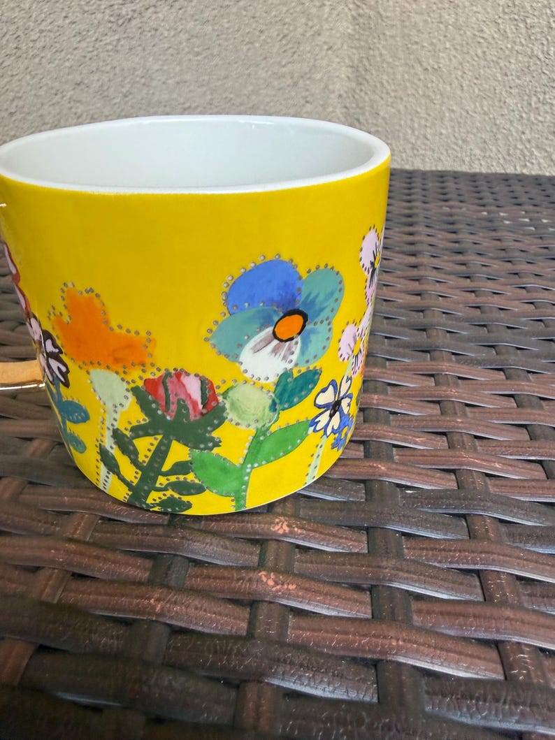 May include: A yellow ceramic mug with a white interior, adorned with a colorful floral design. The flowers include blue, orange, pink, and white blooms with green leaves. The mug is cylindrical and sits on a brown woven surface.